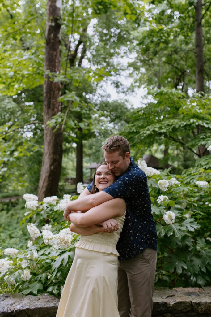 man hugging woman's shoulders from behind in front of flowers