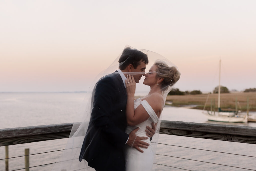 groom hugs bride, while she lightly touches his face in front of a beautiful sunset and sailboat over the water